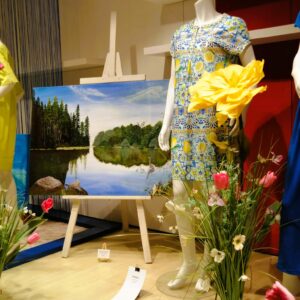 Großformatiges blaues See-Gemälde von Johanna Degelmann inmitten von gelben und blauen Sommerkleidern im Schaufenster vom Modehaus Streit in Ettlingen